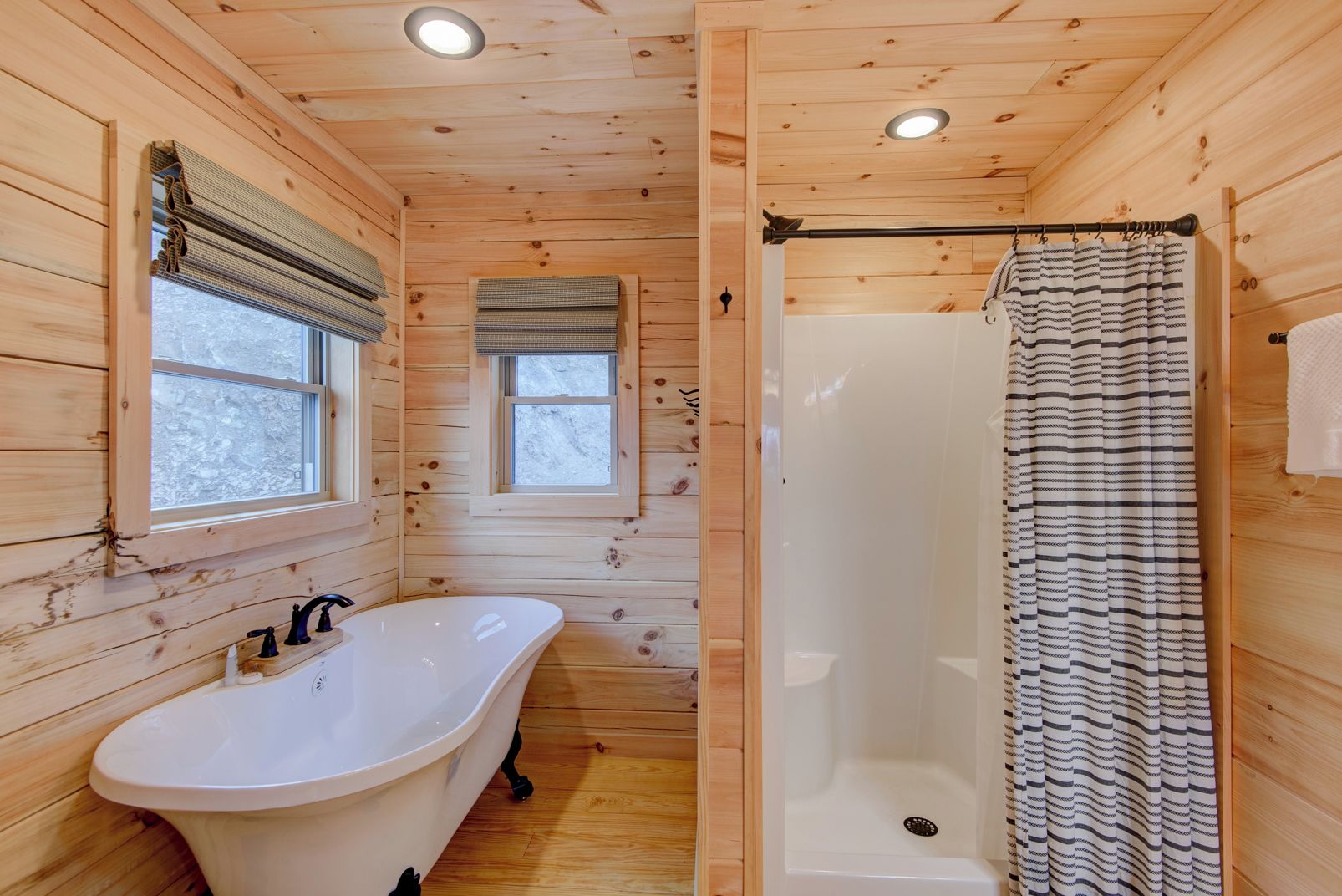 A wooden cabin bathroom with a freestanding bathtub, a shower with a striped curtain, and two windows with blinds.