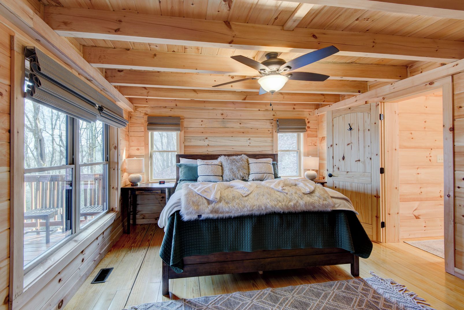 A wooden cabin bedroom with a bed, two windows, a ceiling fan, and a wooden deck outside.