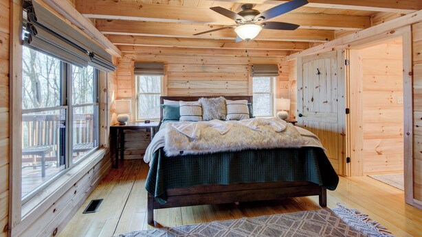 A wooden cabin bedroom with a bed, two windows, a ceiling fan, and a wooden deck outside. - High Time Cabin (Banner Elk)
