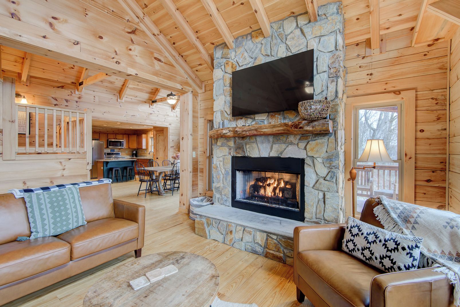 A cozy cabin interior with a stone fireplace, wooden walls, a flat-screen TV, and a dining area.