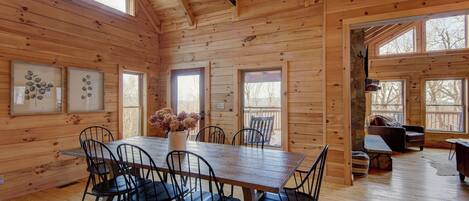 Interior de una cabaña de madera con una mesa de comedor, sillas, un sofá y una chimenea.