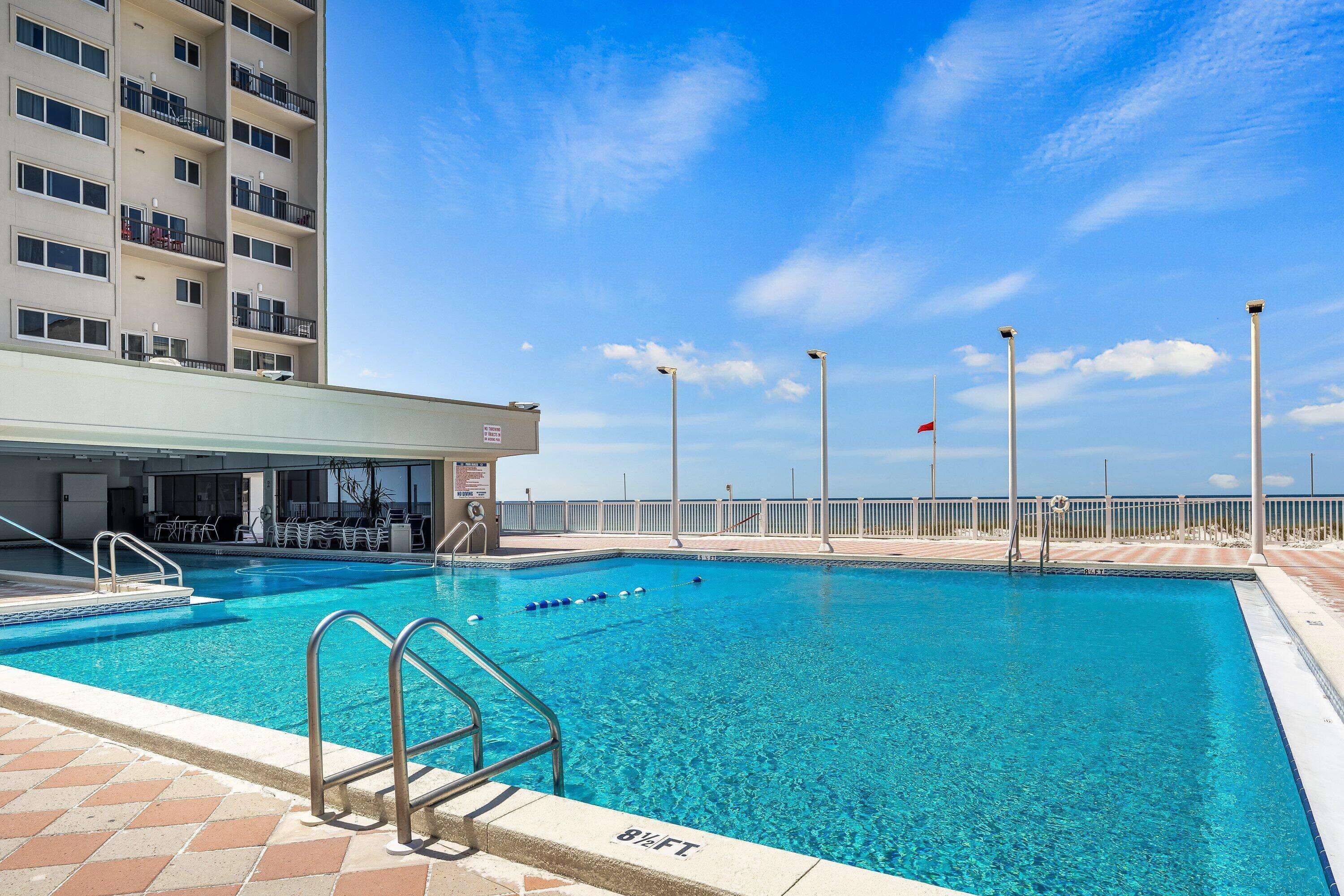A swimming pool with a clear blue water, a covered area with chairs, and a building with balconies in the background.
