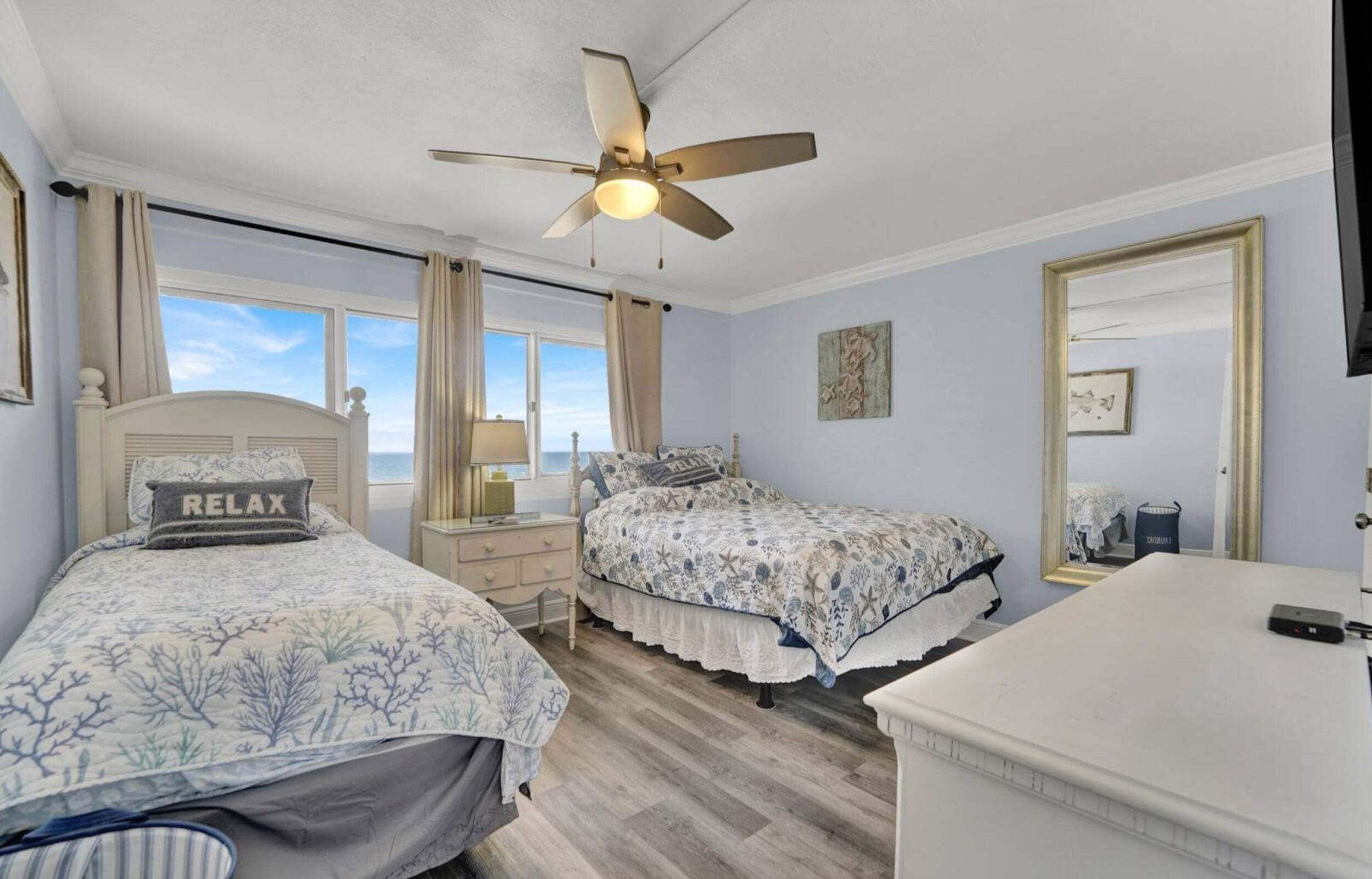 A bedroom with two beds, a ceiling fan, a window with curtains, a bedside table with a lamp, and a dresser.