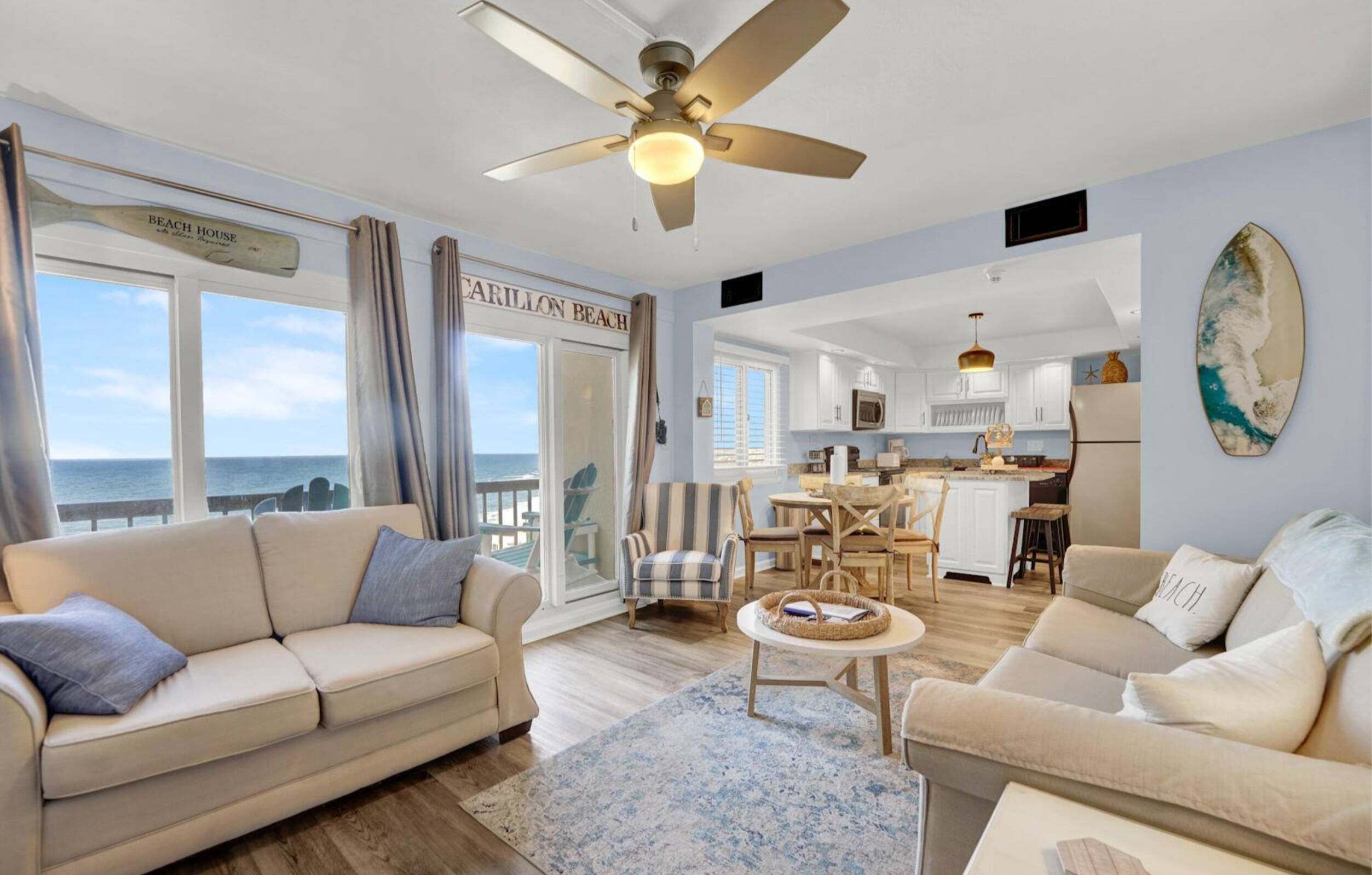 Un salon comprenant un canapé, des fauteuils, une table basse et un ventilateur de plafond. De grandes fenêtres offrent une vue sur l’océan.