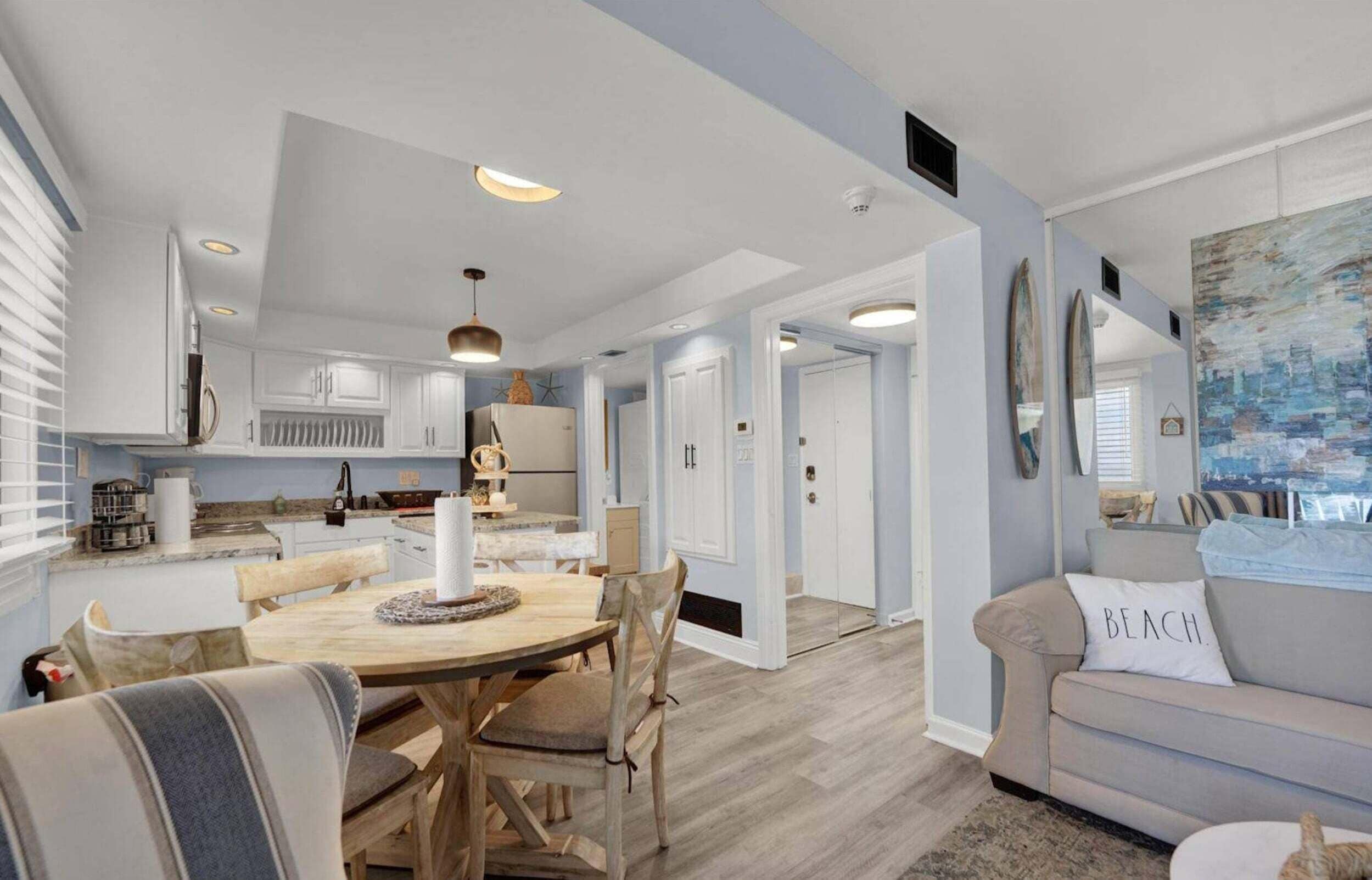 A spacious living area with a dining table, a sofa with a beach-themed pillow, and a kitchen with white cabinets.