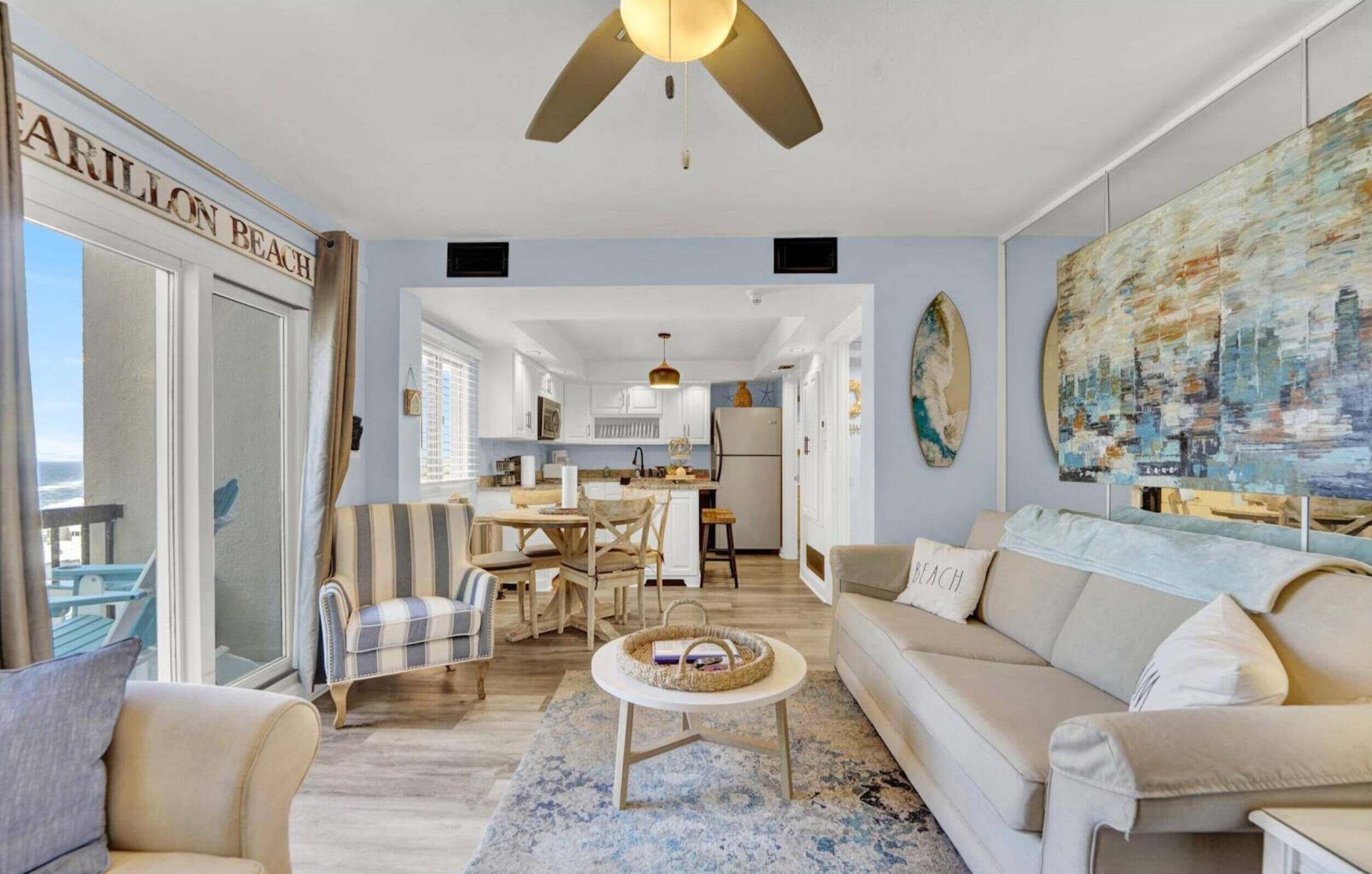 A spacious living room with a large window, a ceiling fan, a dining area, and a kitchen in the background.