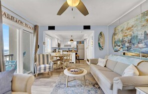 A spacious living room with a large window, a ceiling fan, a dining area, and a kitchen in the background.