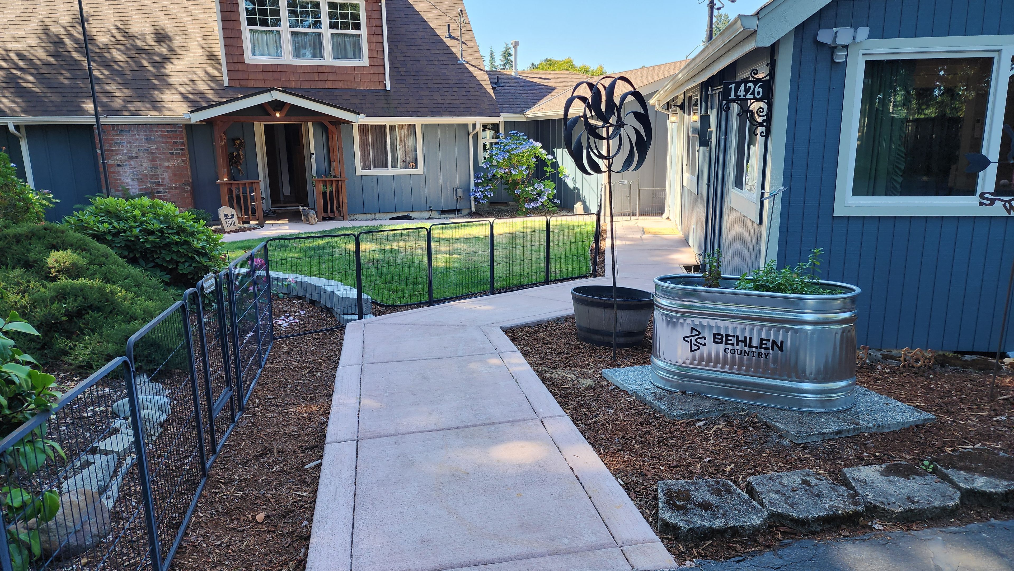 A residential property with a paved driveway, a metal fence, a landscaped garden, and a metal planter with a sign.