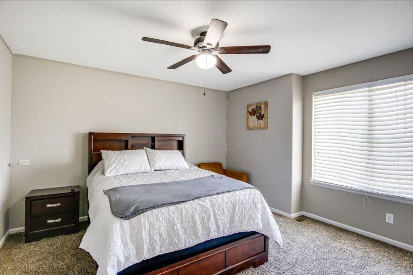 A bedroom with a bed, a nightstand, a ceiling fan, a window with blinds, and a small painting on the wall.