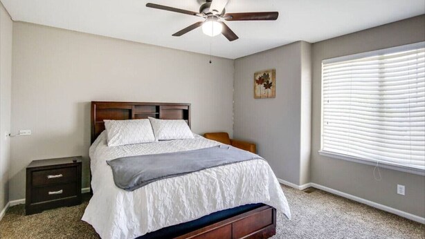 Una camera da letto con un letto, un comodino, una ventola a soffitto, una finestra con persiane e un piccolo quadro appeso al muro.