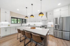 A modern kitchen with white cabinets, a large island with bar stools, a stainless steel refrigerator, and pendant lighting. - Creek Crossing at Eagles Nest (Banner Elk)