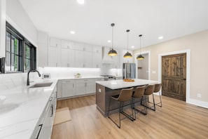 A modern kitchen with white cabinetry, a central island with bar stools, pendant lighting, and a large wooden door. - Creek Crossing at Eagles Nest (Banner Elk)