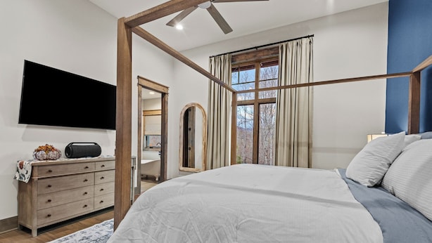 A bedroom with a four-poster bed, a flat-screen TV, a wooden dresser, and a view of trees through the window.