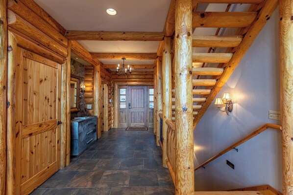 A wooden log cabin interior with a staircase, stone flooring, and a chandelier.