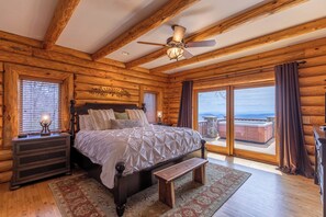 A log cabin bedroom with a large bed, wooden headboard, a nightstand, a bench, and a view of the outside.