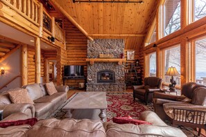 A log cabin interior with a stone fireplace, leather furniture, and large windows.