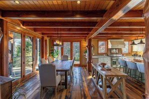 A spacious wooden cabin interior with a large dining table, chairs, a kitchen area, and large windows offering views of the surrounding forest.