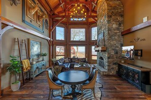 A spacious living room with a stone fireplace, large windows, and a high ceiling with wooden beams. - Bailey's Creek Lodge (Hudson)