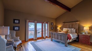 Ein Schlafzimmer mit einem großen Bett, einem hölzernen Kopfteil, einem Nachttisch, einem Sessel und Blick durch die Glastüren.