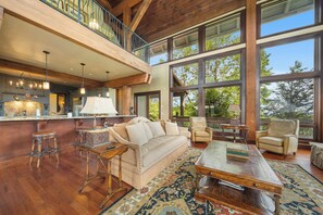 A spacious living area with a high ceiling, large windows, a wooden floor, a sofa, a coffee table, and a bar area. - Overlook at Wilderness Trail (Linville)