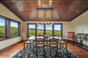 A dining room with a large table, six chairs, a chandelier, and a view of a forest through large windows. - Overlook at Wilderness Trail (Linville)