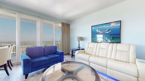 A hotel room with a balcony, a blue sofa, a white sofa, a glass coffee table, and a painting of an airplane. - Laketown Wharf 1738 (Panama City Beach)