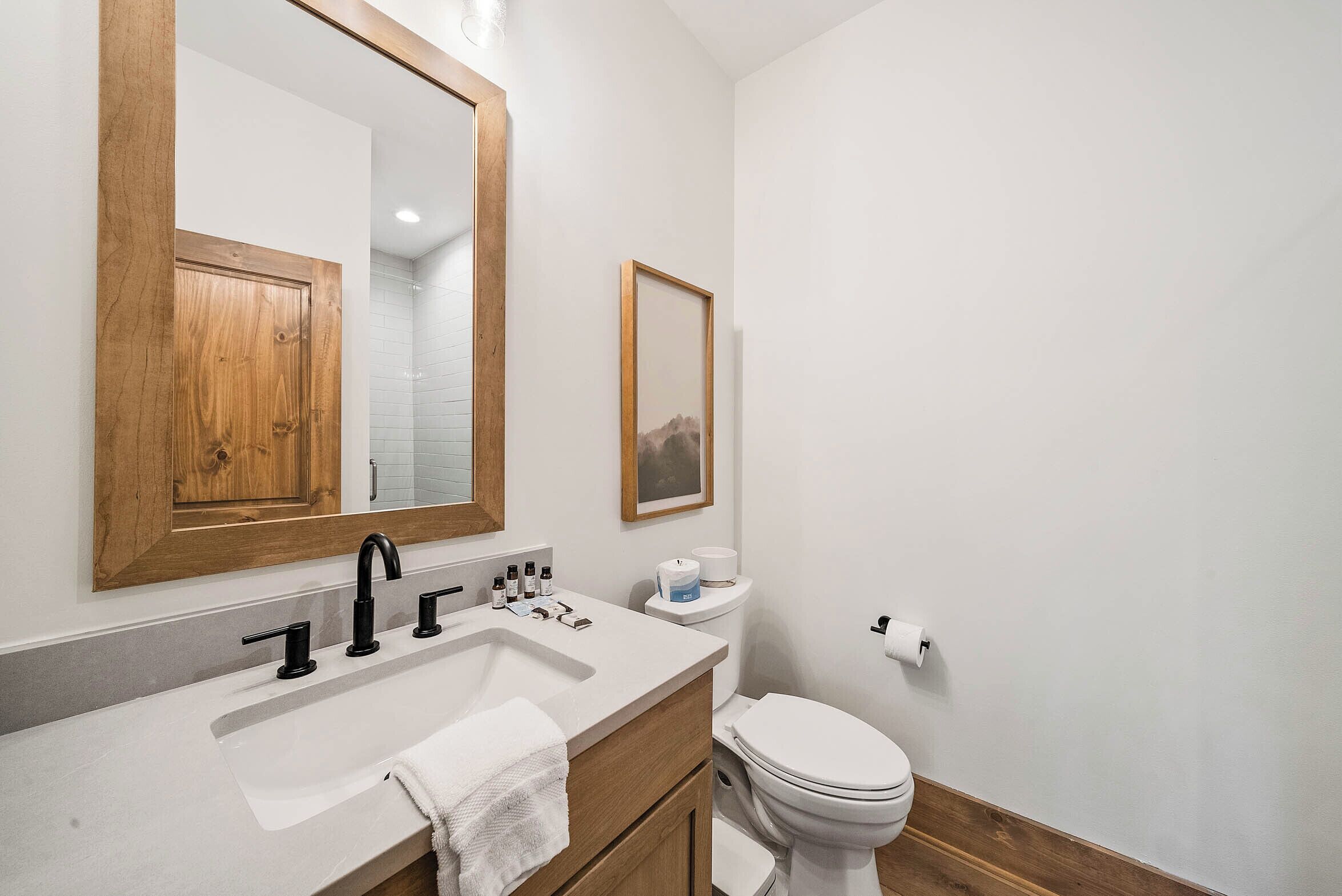 A bathroom with a wooden vanity, a large mirror, a white sink, a toilet, and a framed picture on the wall.