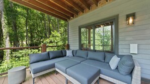 A covered outdoor seating area with a large sectional sofa, a concrete planter, and a view of a forested area. - Boulder Springs Lodge at Eagles Nest (Banner Elk)