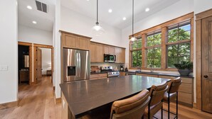 A modern kitchen with wooden cabinets, stainless steel appliances, a large island with bar stools, and large windows. - Boulder Springs Lodge at Eagles Nest (Banner Elk)