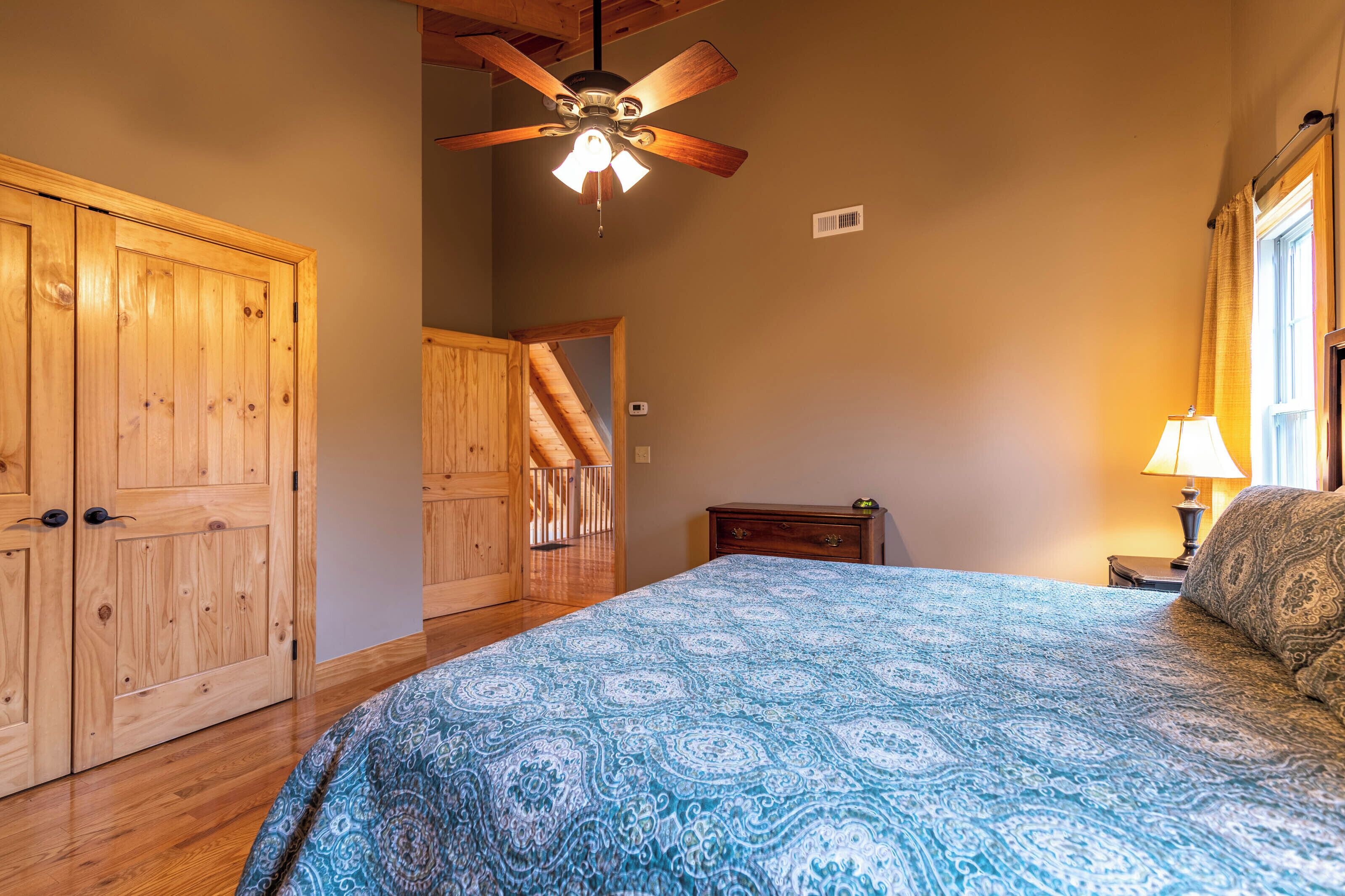 A bedroom with a large bed, wooden wardrobe, ceiling fan, and a nightstand with a lamp.