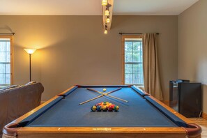Una mesa de billar con bolas y tacos, una lámpara y un televisor en una habitación con ventanas y cortinas.