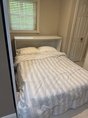 Ein Schlafzimmer mit Bett, einem Fenster mit Jalousien, einer Tür und einem kleinen Regal über dem Bett.