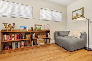 Ein Zimmer mit grauem Sofa, einem Holzregal mit Büchern und gerahmten Bildern sowie einer Stehlampe.