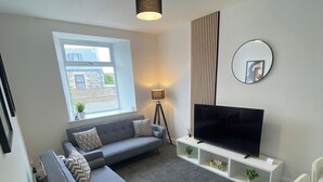 Ett modernt vardagsrum med en grå soffa, en platt-TV, ett litet bord och ett fönster med utsikt över byggnader.