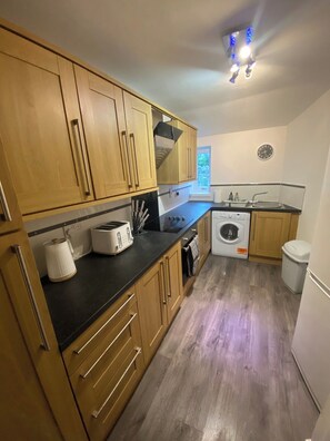A compact kitchen with wooden cabinets, a built-in oven, a washing machine, and a sink. - The Haven at Sandhaven (Sandhaven)
