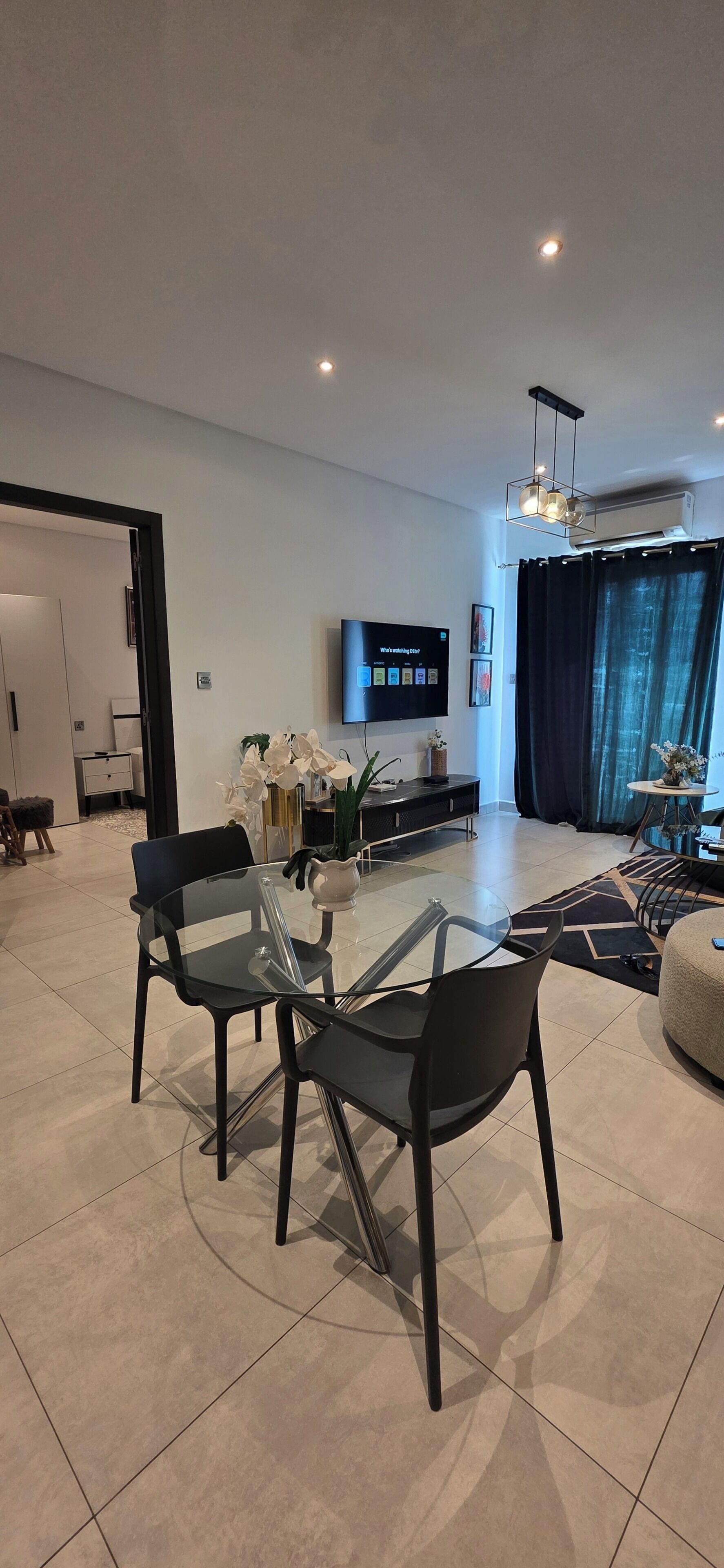 A modern living room with a flat-screen TV, a dining area with glass-top tables, and a large window with curtains.