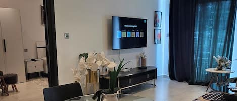 A modern living room with a flat-screen TV, a dining area with glass-top tables, and a large window with curtains.