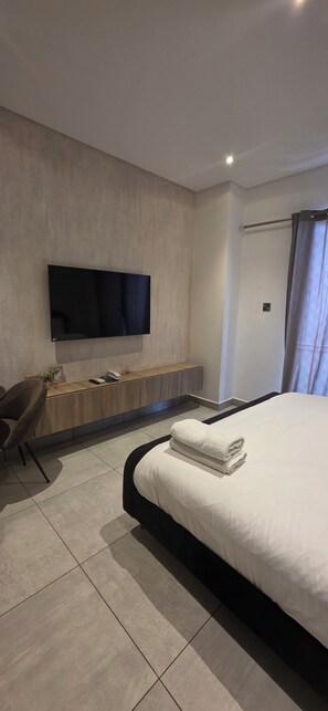 A modern bedroom with a large bed, a flat-screen TV mounted on the wall, a wooden bench, and a chair. - signature Apartments Exquisite (Accra)