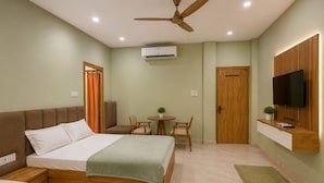 A hotel room with two beds, a ceiling fan, an air conditioning unit, a TV, a small table with chairs, and a wooden door.
