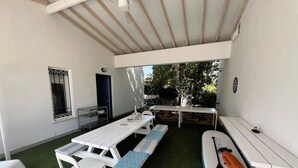 Een overdekt terras met een tafel en banken, een bijzettafeltje met een stoel, en een surfboard.