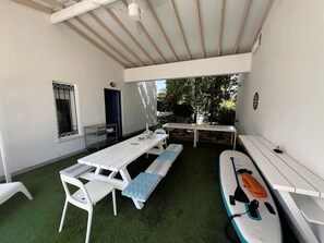 A covered patio with a table and benches, a small table with a chair, and a surfboard. - Protaras Cyprus Beach House (Paralimni)