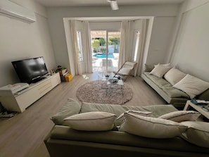 A modern living room with a sofa, TV, and a view of a swimming pool. - Protaras Cyprus Beach House (Paralimni)
