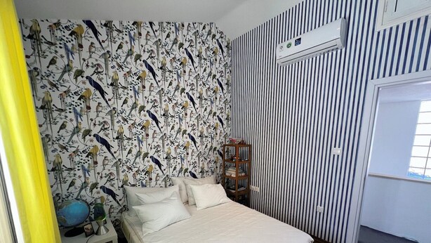 Een slaapkamer met een bed, nachtkastje, lamp, boekenplank en een wandairconditioner.