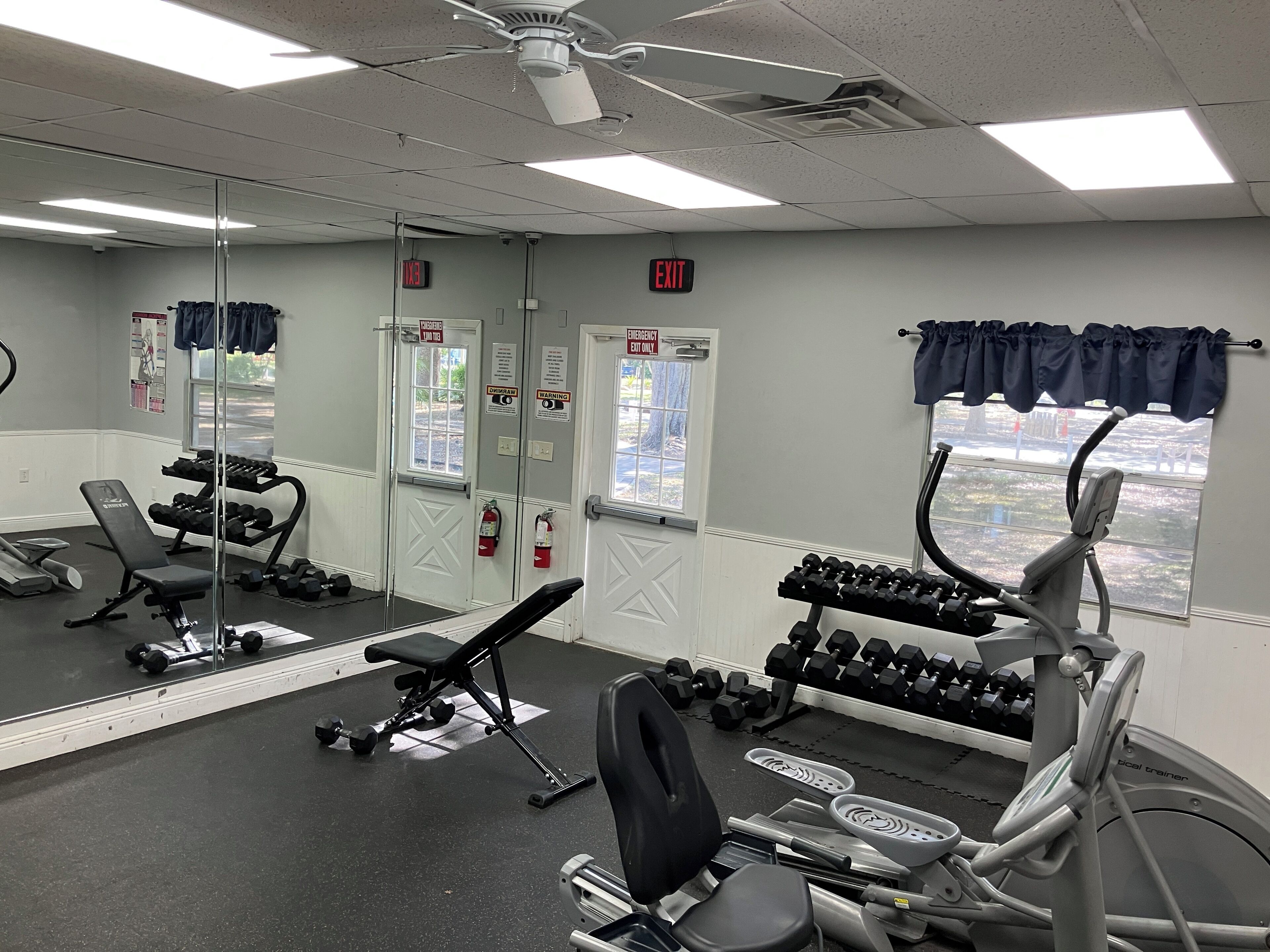 Un gimnasio con equipo de ejercicio, un espejo y señales de salida de emergencia.
