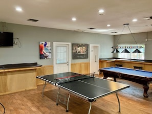 A billiards room with a pool table, a ping pong table, and a bar area.