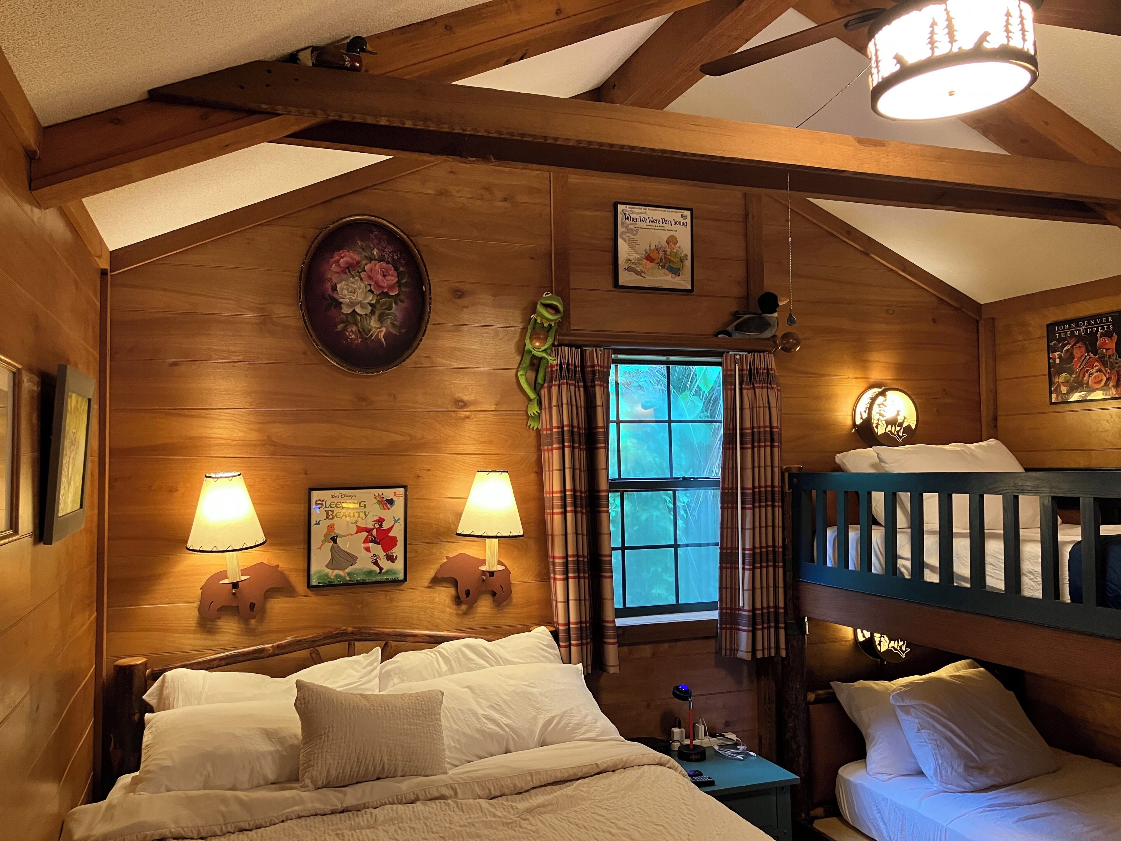 A wooden cabin room with a bunk bed, two wall-mounted lamps, a window with plaid curtains, and a poster of 'Sleeping Beauty'.