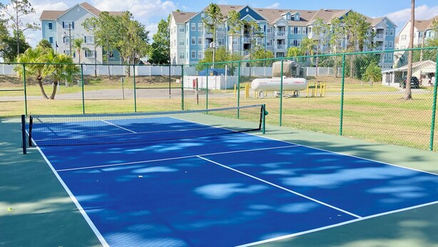 A tennis court with a net, surrounded by a green fence, and residential buildings in the background.