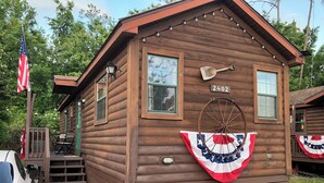 A wooden cabin with a number '2402' on the front, a porch with a bench, and an American flag.