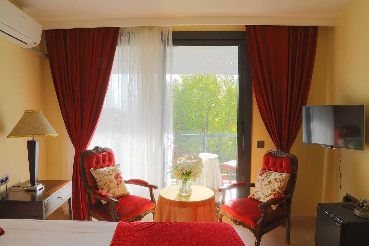 A hotel room with two red upholstered chairs, a small round table with a vase, a bed, a TV, and an air conditioning unit.
