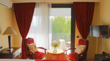 A hotel room with two red upholstered chairs, a small round table with a vase, a bed, a TV, and an air conditioning unit.